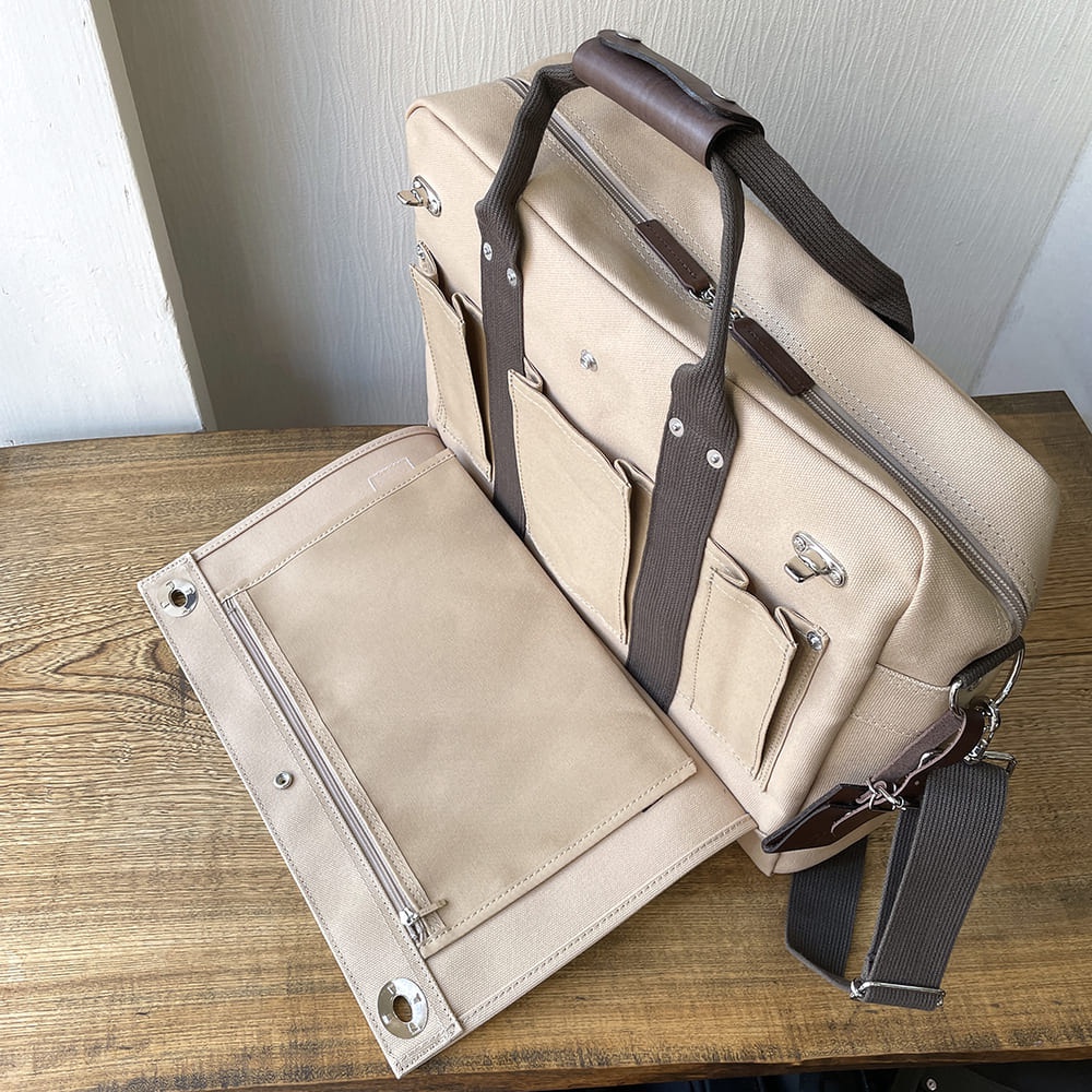 米色純綿帆布製成的商務公事包，設有深棕色皮革提把和織帶，包身外側有數個口袋，附有可拆卸的肩帶和一個可打開的隔層。