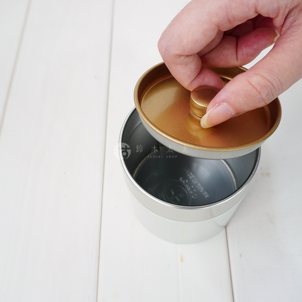 簡約錫製茶葉罐，罐身為銀白色，罐蓋為金色，帶有圓形提鈕。一只手正將罐蓋從罐口上方移開。
