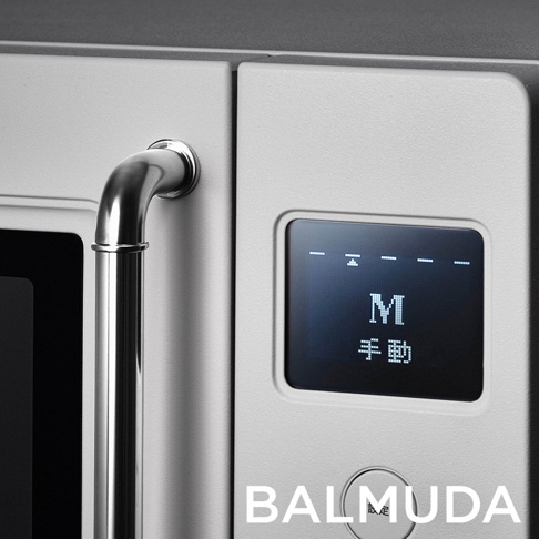 BALMUDA 微波爐的近距離細節特寫，畫面呈現帶有霧面金屬質感的淺灰色機身，左側配備金屬圓弧把手，右側為顯示面板與控制旋鈕，數位螢幕正顯示英文字母 M 以及日文手動二字，整體設計簡約且極具工業風格。