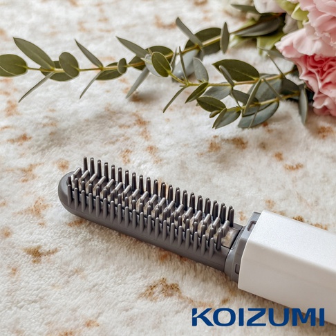 灰色的迷你瀏海梳，梳齒密集，手把為白色，背景有綠葉和粉色花朵裝飾，右下角有KOIZUMI品牌標誌