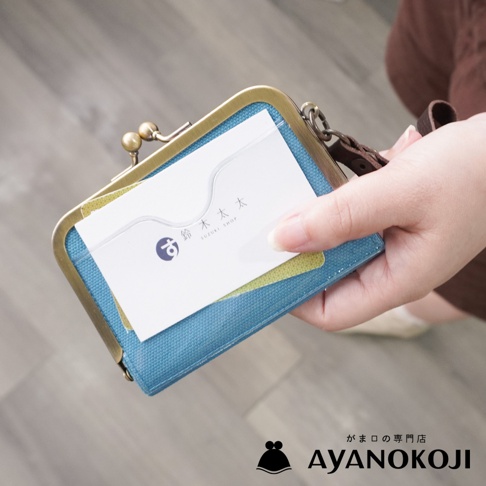 一個手拿著 AYANOKOJI 品牌京都職人手作帆布橫式口金卡片收納包，包包是藍綠色，裡面放著一張名片。