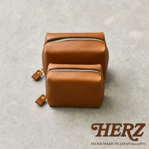 兩款大小不同的職人手作皮革箱型收納包，皆為棕色，搭配金色拉鍊與同色皮革拉環，擺放在灰色紋理背景上，下方有HERZ品牌標誌與HAND MADE IN JAPAN Since1973字樣