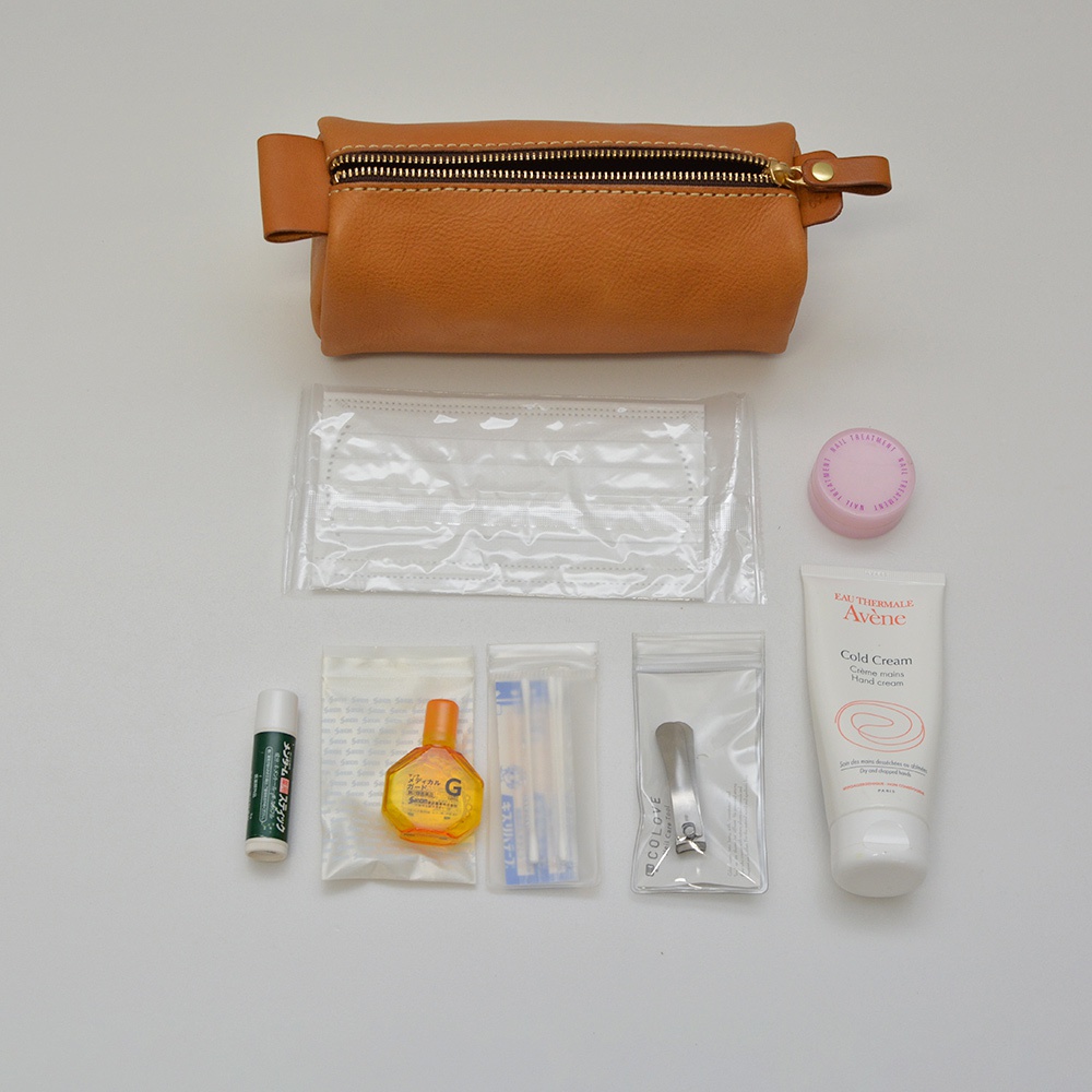 職人手作皮革多用途收納筆袋，皮革顏色為棕褐色，配有金色拉鍊。袋內展示有口罩、護手霜、眼藥水、指甲剪、護唇膏及其他小物件。