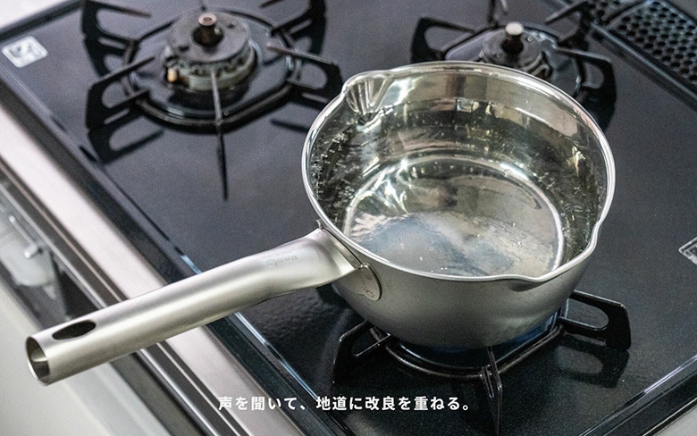 一個裝有水的銀色不鏽鋼鍋，帶有長柄，放在黑色爐灶上。鍋的內壁有刻度線，鍋緣有傾倒口。