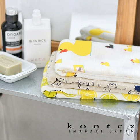 折疊堆放的 KONTEX 輕薄透氣萬用浴巾，頂層印有可愛的小黃鴨圖案，中間層為簡約的魚骨線條設計，底層則鋪滿明亮的檸檬切片圖樣。旁邊擺放著白色的沐浴乳瓶與一塊肥皂，整體呈現溫馨舒適的居家沐浴氛圍。