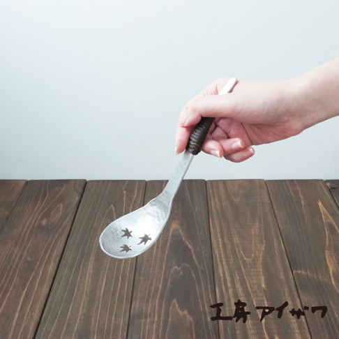 一隻手正拿著一把鋁製中型瀝水湯勺，湯勺表面帶有錘目紋，勺面上有三個楓葉形狀的鏤空孔。湯勺的握柄處纏繞著深棕色的繩索。