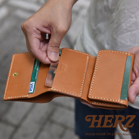 一雙手正拿著一個日本HERZ手工製作的淺棕色皮革三折迷你短夾，皮夾內部結構包含紙鈔夾層與多個卡片收納槽，正展現出打開後的實用配置，皮革表面刻有HERZ品牌標誌，整體設計展現細膩的職人縫線質感。
