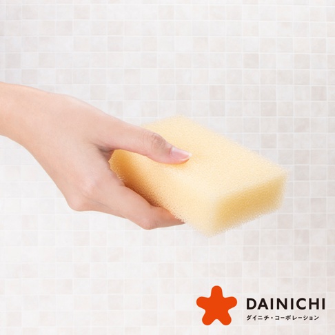 一隻手拿著一塊淺黃色的海綿，海綿呈長方形，材質為多孔泡沫。背景是淺色馬賽克瓷磚。