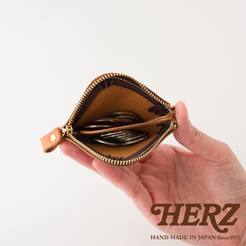 一隻手拿著日本HERZ職人手作皮革零錢包，拉鍊敞開呈現內部結構，可以看到棕色皮革內裡分為兩個隔層，裝有少量硬幣。背景為素面純白，右下角印有HERZ品牌標誌與產地說明。
