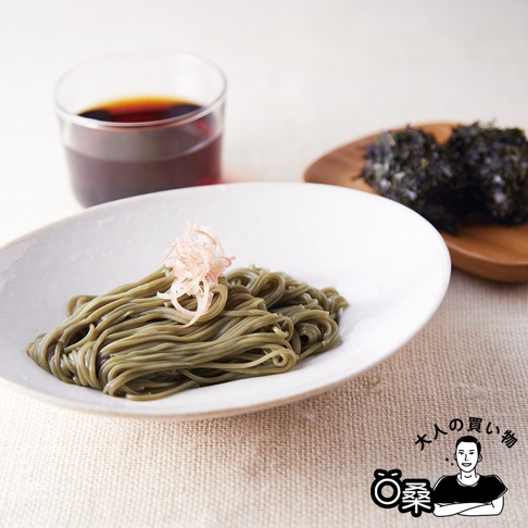 一盤綠色蕎麥麵，上方點綴著粉紅色薑絲，旁邊有一個裝有深褐色醬汁的玻璃杯，以及一塊木盤上擺放著兩團海苔。