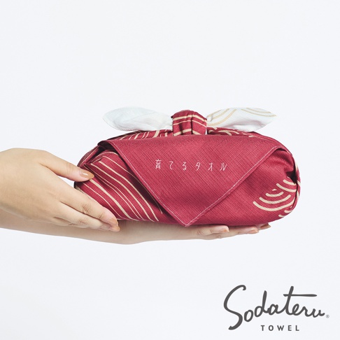 一雙手捧著以紅色布料包裹的浴巾，布料上有著白色和金色的條紋及水波紋圖案，布料上方打成了蝴蝶結的形狀，中間繡有日文「育てタオル」字樣，下方角落印有「Sodateru TOWEL」的品牌標誌。