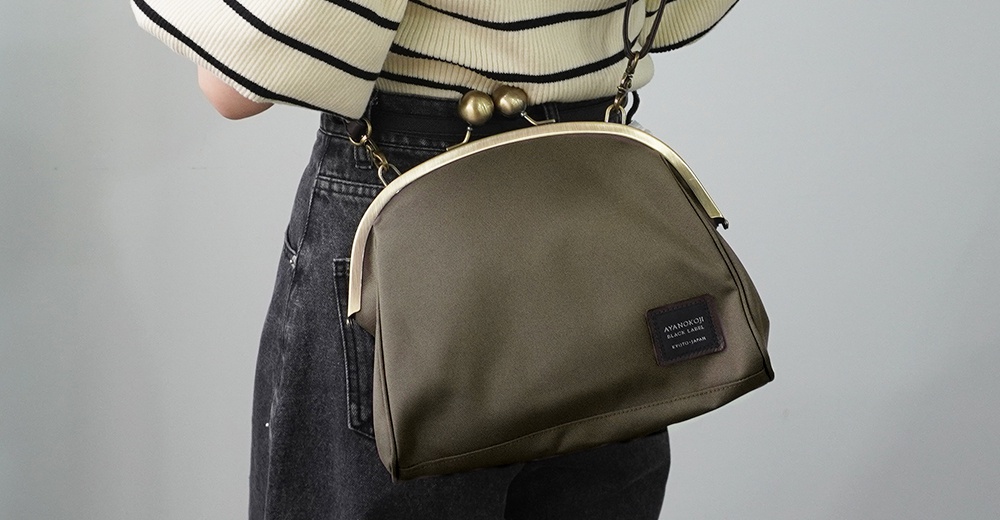 斜背包為軍綠色，採用CORDURA®材質，包上有 AYANOKOJI BLACK LABEL KYOTO-JAPAN 字樣的織標，背帶為古銅金色的球形金屬扣環