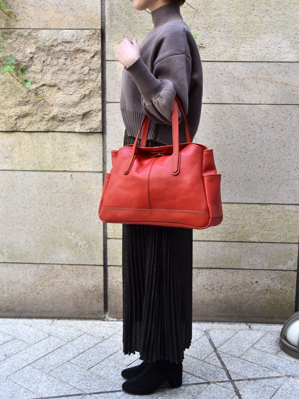 一位女性身穿灰色高領毛衣和黑色百褶裙，肩上斜背著一個亮紅色皮革橫式托特包，包包有兩個手提帶和側邊口袋。