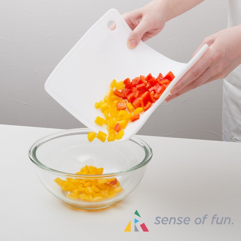 白色矽膠砧板，傾斜著將切塊的紅、黃甜椒倒入玻璃碗中。砧板左上角有一個圓形掛孔。
