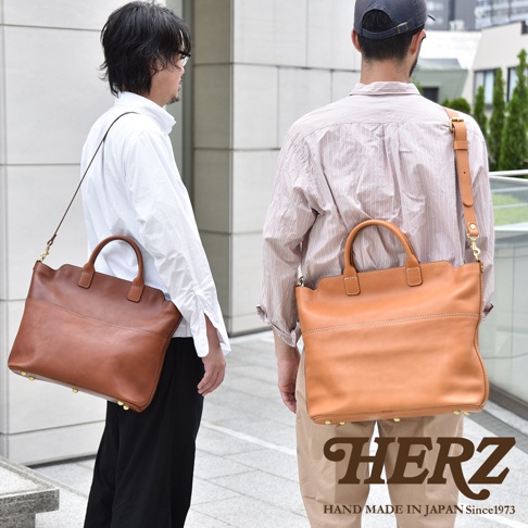 兩位男士各背一個職人手作皮革兩用托特包，一個為棕褐色，一個為淺棕色。包款為方形，有提把和可拆卸肩帶，底部有四個金屬腳釘。