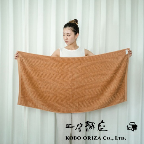 一位女性手持一條淺棕色人字紋有機棉浴巾，浴巾紋理清晰。