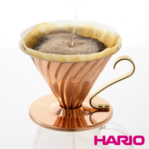 V60銅製濾杯，帶有螺旋紋路，呈現溫潤的銅色，搭配金色手柄，濾紙內有咖啡粉和正在注入的水。