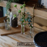 黃銅可夾式玻璃花器，兩個玻璃試管狀花瓶，分別插入綠色花苞和尤加利葉，由黃銅夾子固定在木質層架上。