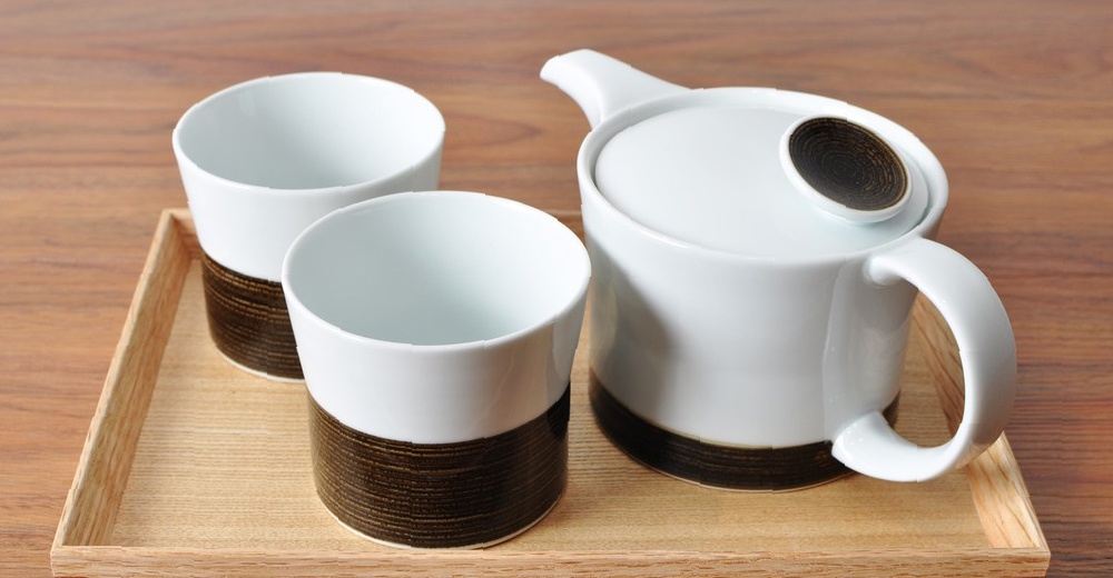 白瓷茶具組，包含一個白色茶壺和兩個白色茶杯，皆帶有深棕色麻紋飾帶。產品置於木紋托盤上。