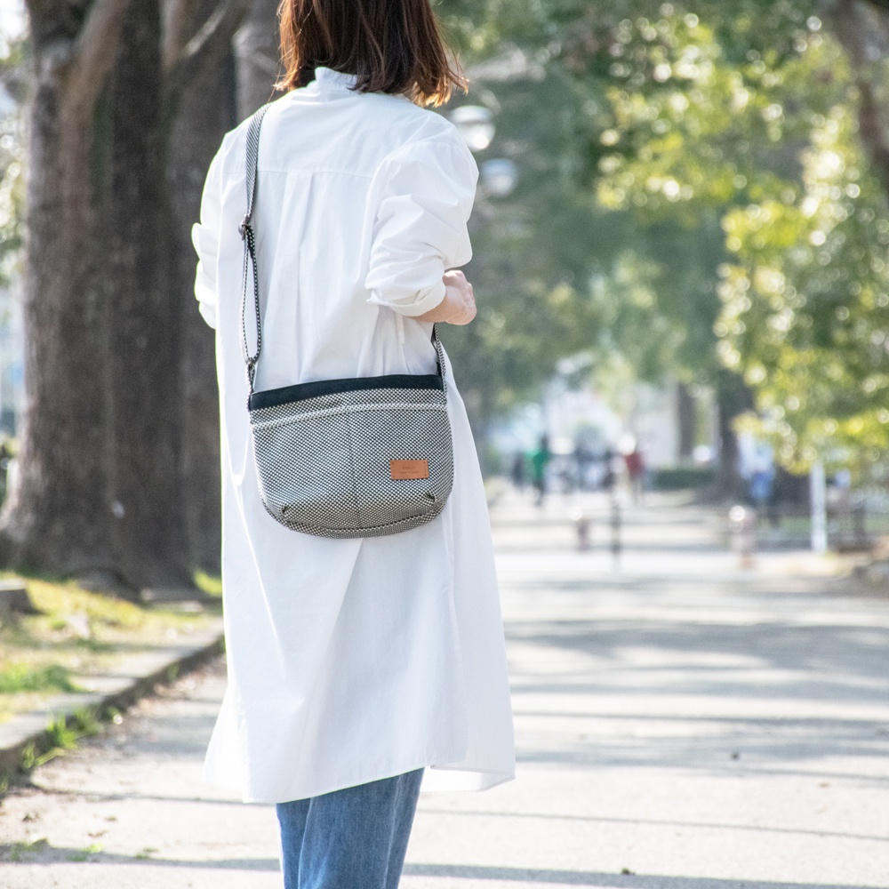 三河木綿刺子織肩背包，採用黑白菱格紋圖案，搭配黑色織帶和皮革標籤，斜挎於身穿白色長版上衣的女性身上。