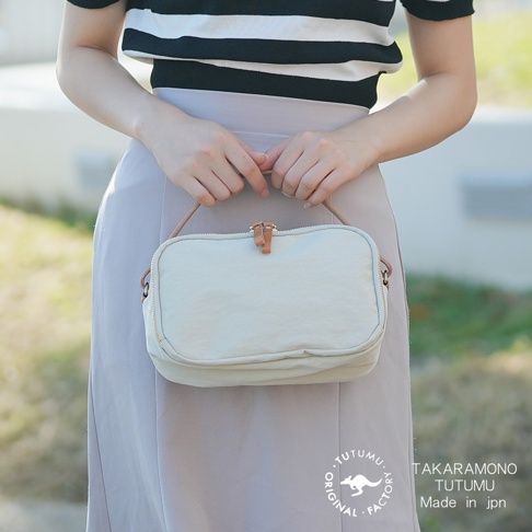 一位女士身穿黑白條紋上衣與灰色裙子，雙手提著一個米白色的手提包，包包上有皮革飾條和拉鍊。