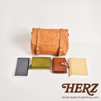 職人手作皮革水洗加工復古郵差包，配有兩個皮帶扣和皮帶。下方展示一個黑色手機、一個橄欖綠色皮革錢包、一個棕色皮革錢包和一個米色筆記本。