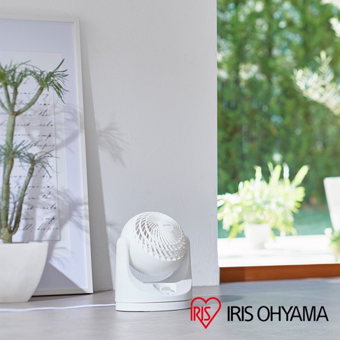 白色IRIS OHYAMA PCF-MKM15空氣循環扇，具有可調節角度的圓形扇頭和簡潔的底座，放置在室內地板上，背景有綠色植物和窗戶。
