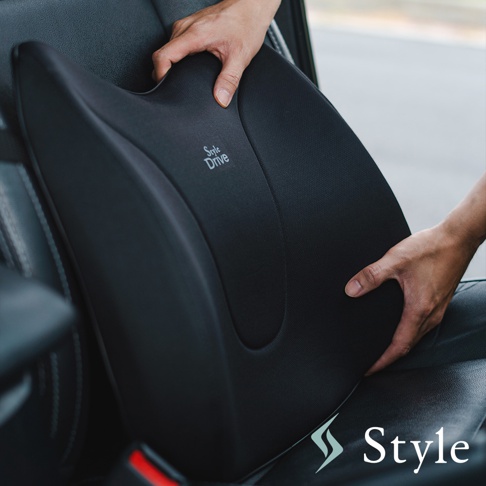 黑色織物材質的 Drive S 車用舒適靠腰椅，具有人體工學曲線，表面印有白色「Style Drive」字樣。兩隻手正將靠腰椅放在汽車座椅上。