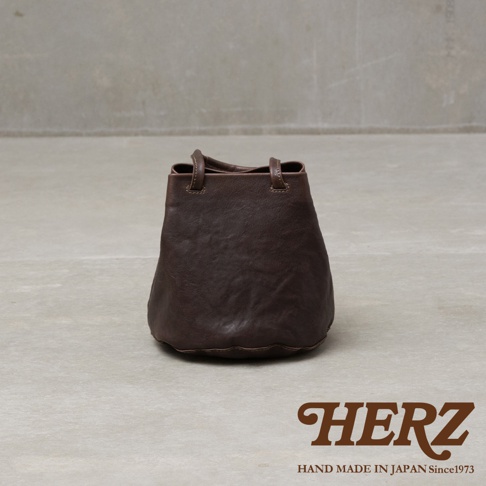 一款由日本HERZ品牌推出的深褐色皮革手提托特包，包身採用柔軟具質感的皮革材質，呈現自然的皺褶紋理。該包款設計簡約，採用抽繩開口樣式並配有兩條纖細的皮革提把，整體以純色調呈現，放置於淺灰色背景中央，右下角印有HERZ品牌標誌及手工製作年份說明。