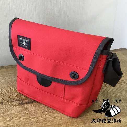 紅色純綿帆布雙扣郵差包，配有黑色滾邊、黑色扣子和黑色肩帶。包的正面有一個黑色皮革裝飾片，上面有一個黑犬印製作品所的黑色標籤。