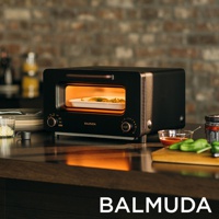黑色 BALMUDA The Toaster Pro 蒸氣烤麵包機，內部有裝著食物的白色烤盤，烤箱內發出溫暖的橘色光芒。產品設有旋鈕和顯示溫度與模式的標示。
