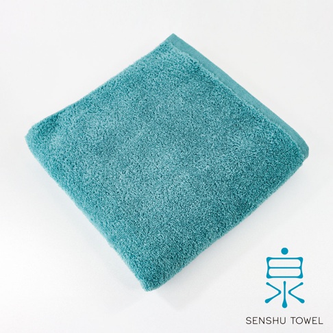 一張藍綠色長纖浴巾，材質蓬鬆柔軟，邊緣有縫線，左上角為浴巾一角，右下角有日文「泉」字樣與 SENSYU TOWEL 商標。