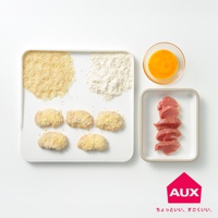 白色砧板上擺放著麵包糠、麵粉、裹好麵包糠的肉塊，旁邊有一個裝有蛋液的玻璃碗和一個裝有切片生肉的白色長方形盤。