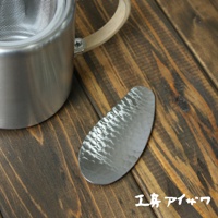 一個橢圓形的不鏽鋼茶勺，表面有槌紋紋理，旁邊是一個不鏽鋼茶壺，配有木質手柄。