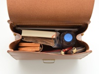 職人手作皮革屋瓦風兩用單肩包 (CW-3)，內含一本淺色書、一本筆記本、一個橘色皮夾、一個深棕色拉鍊包、一個紅色零錢包、一瓶藍蓋礦泉水和一條藍白格紋布。包款為棕色皮革材質，開口向上，可見內部結構。