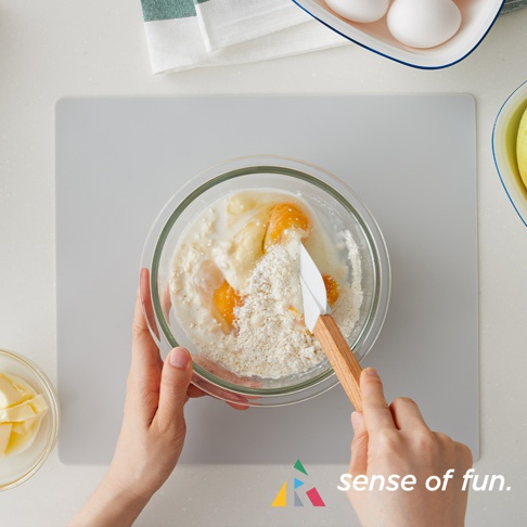 淺灰色矽膠防污墊，上面放著混合雞蛋、牛奶和麵粉的玻璃碗，以及一個木柄矽膠刮刀。