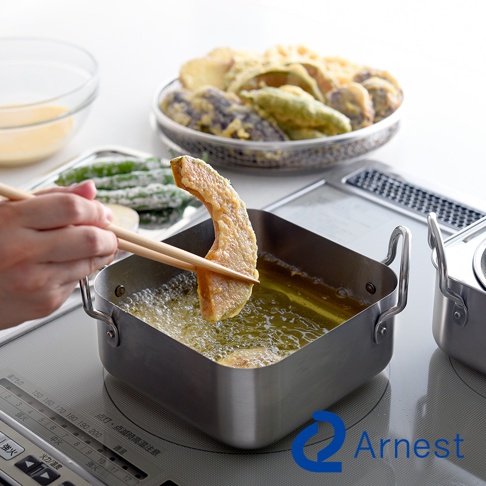 銀色不鏽鋼方型炸物鍋，鍋內裝有熱油，一塊裹粉的南瓜片正被木筷夾起準備油炸。背景有濾油網盛裝炸好的天婦羅，以及裝有麵糊的玻璃碗。