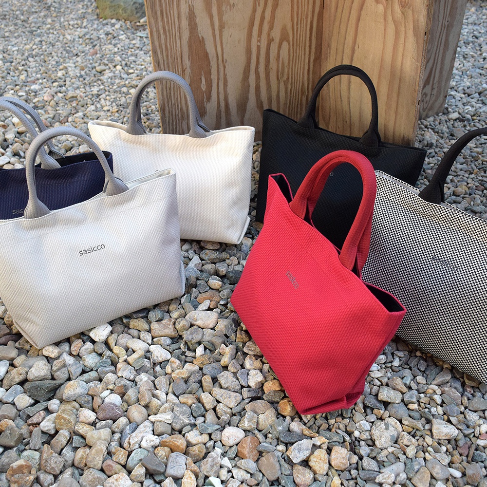 五款三河木綿刺子織經典托特包，分別為深藍、淺灰、白色、紅色及黑白格紋，皆配有同色系手把，置於碎石及木板背景上。