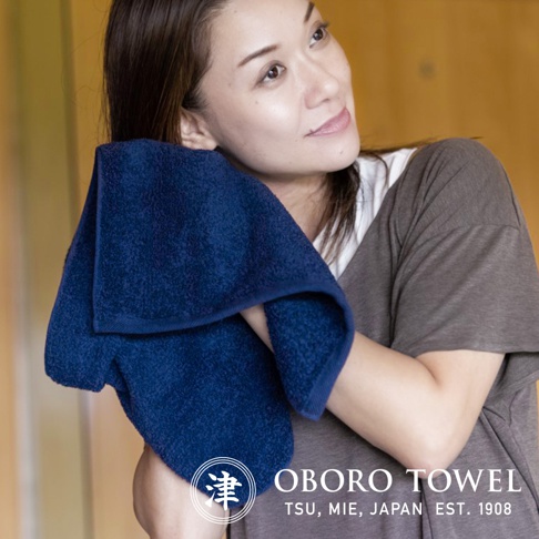 一名女子手持深藍色毛巾，擦拭臉頰。毛巾材質柔軟，邊緣有縫線。背景為淺黃色。