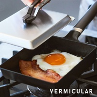 黑色琺瑯鑄鐵玉子燒平底鍋，鍋內煎有太陽蛋和培根，一隻手正用金屬夾子提起配套的銀色鍋蓋。