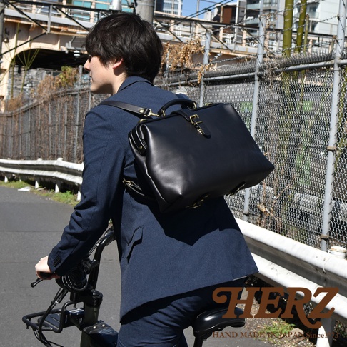 職人手作黑色皮革軟式杜勒斯包，男士著深藍色西裝騎乘自行車，包包以雙肩背方式掛在背後，配有金色金屬扣環。