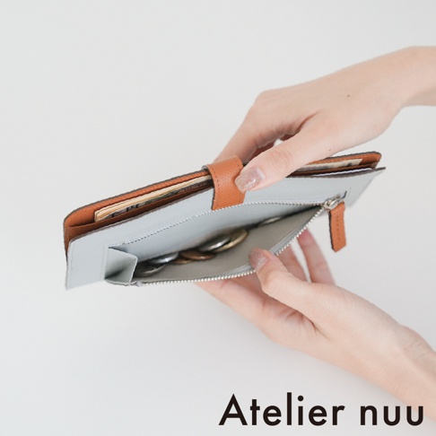 雙手拿著一個打開的 Atelier nuu 雙折長夾，外側為棕色皮革，內側搭配淺灰藍色皮革。長夾中央設有拉鍊零錢袋，內裝有一些硬幣，上方隔層則收納著幾張鈔票，整體設計簡約精緻。