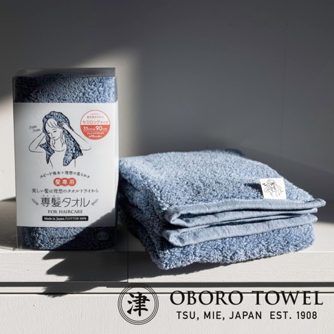 藍色五倍吸水專髮擦髮巾，材質為棉，觸感柔軟，尺寸為 33x90公分，包裝盒上有女性使用擦髮巾的插畫，旁邊疊放著兩條同款擦髮巾。