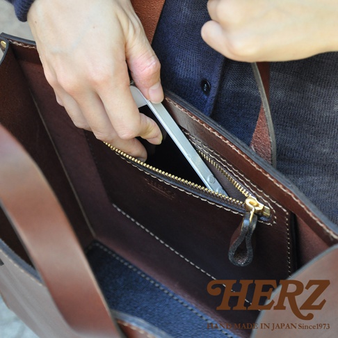 職人手作皮革直式托特包，深棕色皮革，內部有拉鍊口袋，一手正在將手機放入。