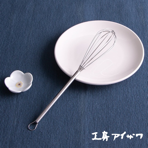 銀色不鏽鋼材質的攪拌棒，帶有圓形握柄環，置於白色圓盤上，旁邊有一個白色小碟，碟子上有花朵圖案，整體擺放在藍色紋理布墊上。