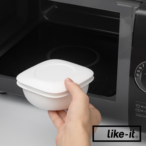 一隻手正將白色簡約雙層蒸氣循環保鮮盒放入微波爐內。保鮮盒為方形設計，帶有圓形蓋子，材質為塑膠。