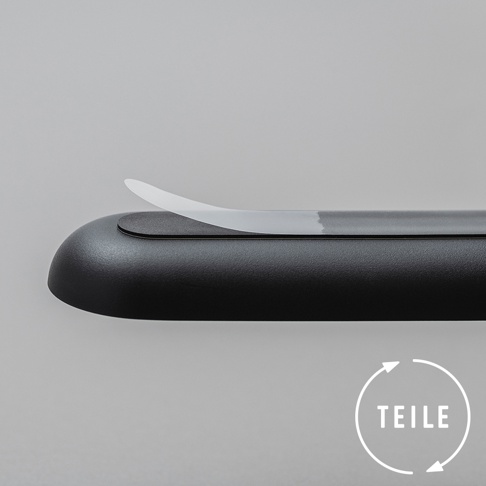 黑色圓潤的產品，表面有細微紋理，上方有一條半透明的白色薄片，部分區域有淺灰色液體痕跡。右下角有一個白色圓圈，內有箭頭循環符號和德語單詞「TEILE」。