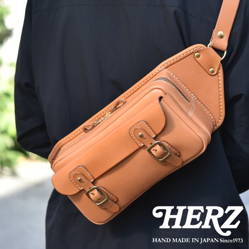 棕褐色皮革製的職人手作斜背包，斜跨於身穿黑衣者身上，包身帶有拉鍊、雙扣環及縫線細節，質感樸實。