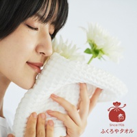 一名女子閉著眼睛，將一條紋理清晰的白色毛巾靠近臉頰，她似乎在感受毛巾的觸感與氣味。毛巾的織紋呈現小方格狀，質地柔軟。背景有兩朵盛開的白色花朵，增添了清新自然的氛圍。右下角有一個紅色的日式圖案標誌，標示著Since1926和日文文字。