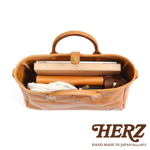 職人手作淺棕色皮革托特包，內部裝有雜誌、錢包和白色布料。包包採用全皮革製作，配有頂部提把和金屬扣環。